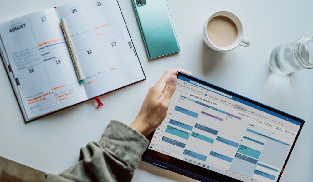 Hand holding tablet with a calendar on it, planner on desk, phone and coffee mug